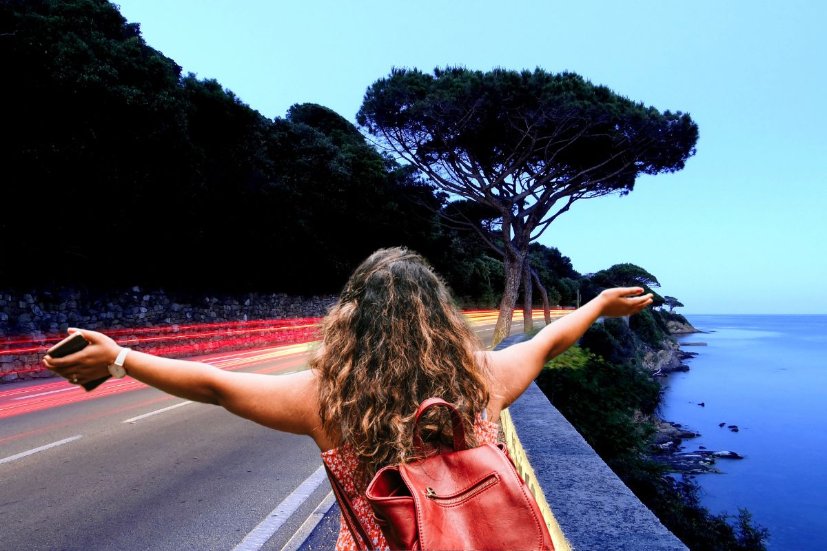 Une femme avec un sac à dos rouge, les bras écartés, sur une route côtière à Sainte Maxime au crépuscule.