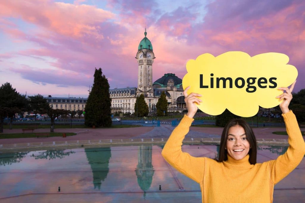 Une femme souriante tenant une pancarte Limoges devant la gare de Limoges au coucher du soleil
