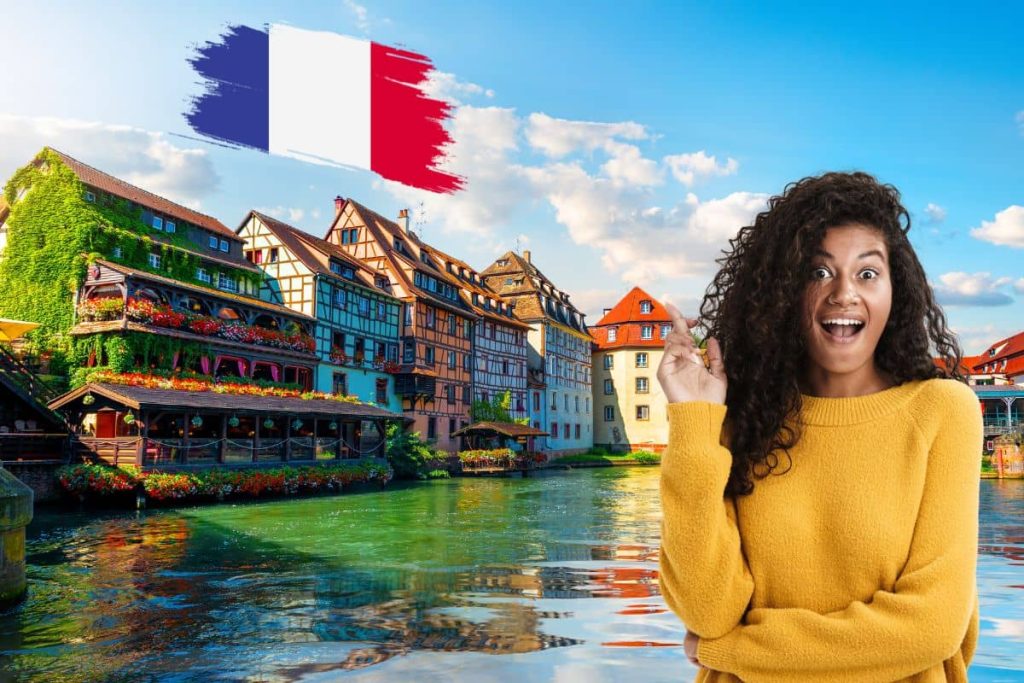 Femme souriante devant des maisons à colombages en France avec un drapeau français en arrière-plan.